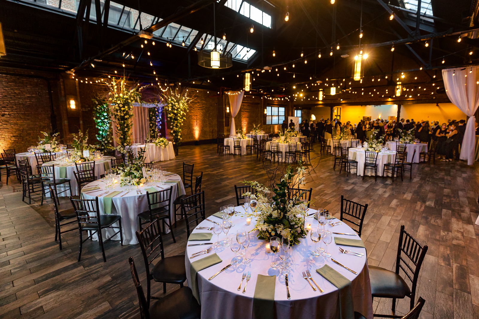 Reception setup at 26 Bridge with round tables, soft candlelight, lush florals, and warm string lights in an industrial loft setting.