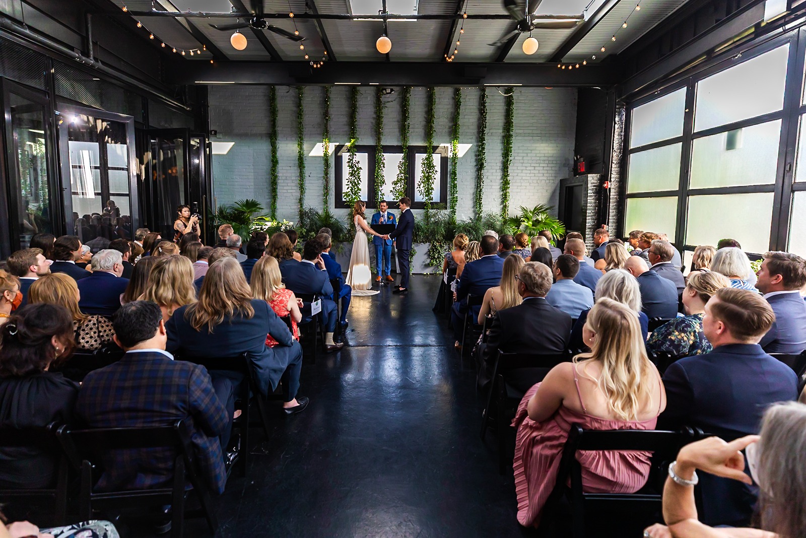 Indoor wedding ceremony at 501 Union with guests seated as couple exchanges vows beneath greenery and soft string lights.
