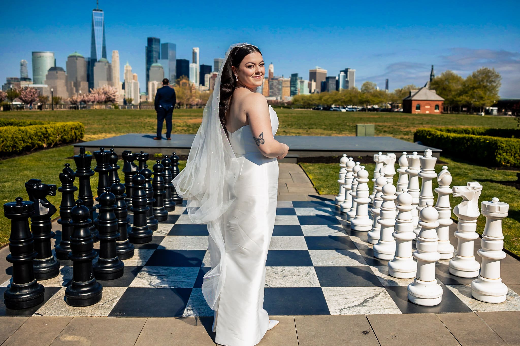 Bride in white dress standing on a giant chessboard with New York City skyline in the background, groom waiting in the distance