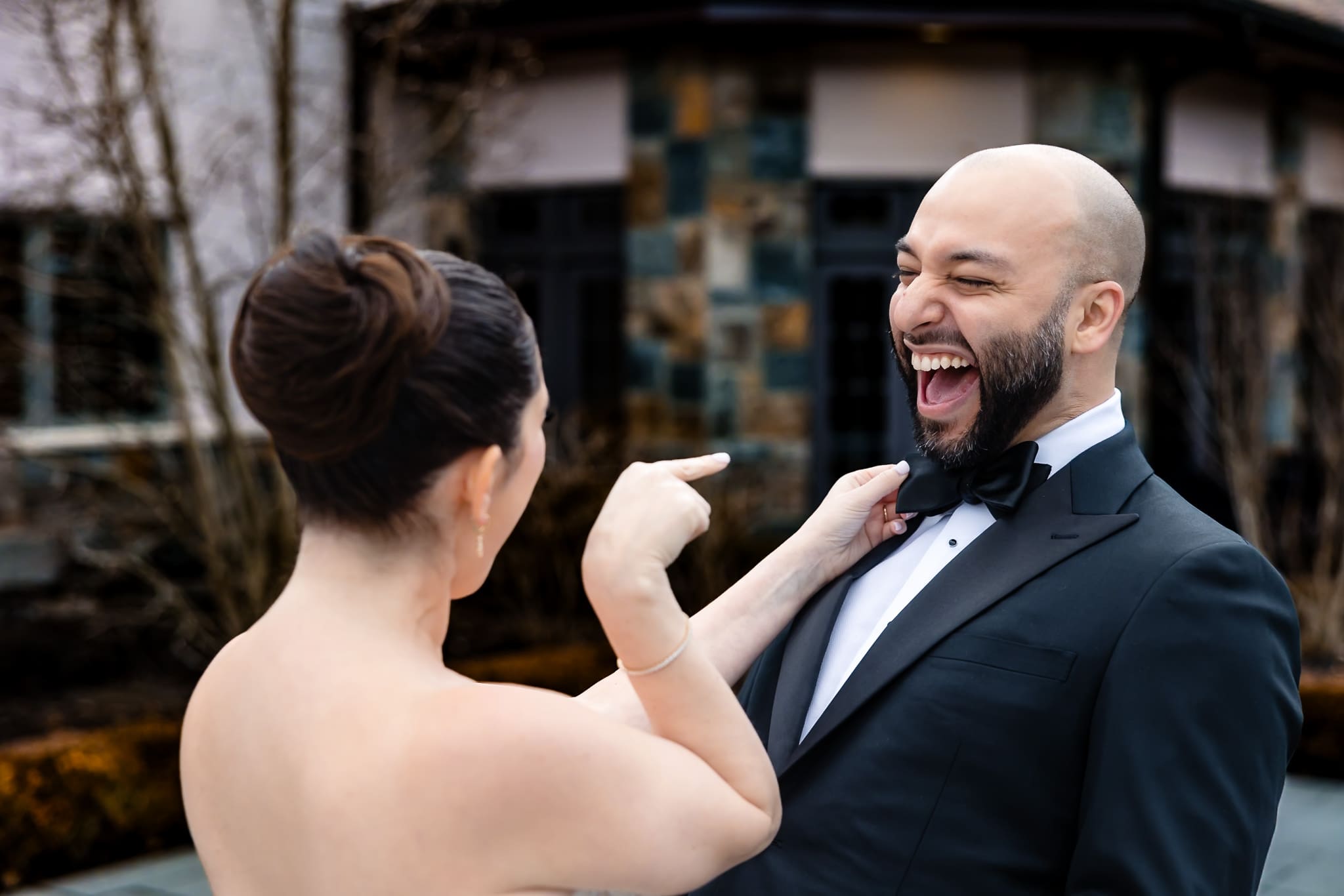 Bride playfully adjusts groom’s bow tie as he laughs during candid wedding moment