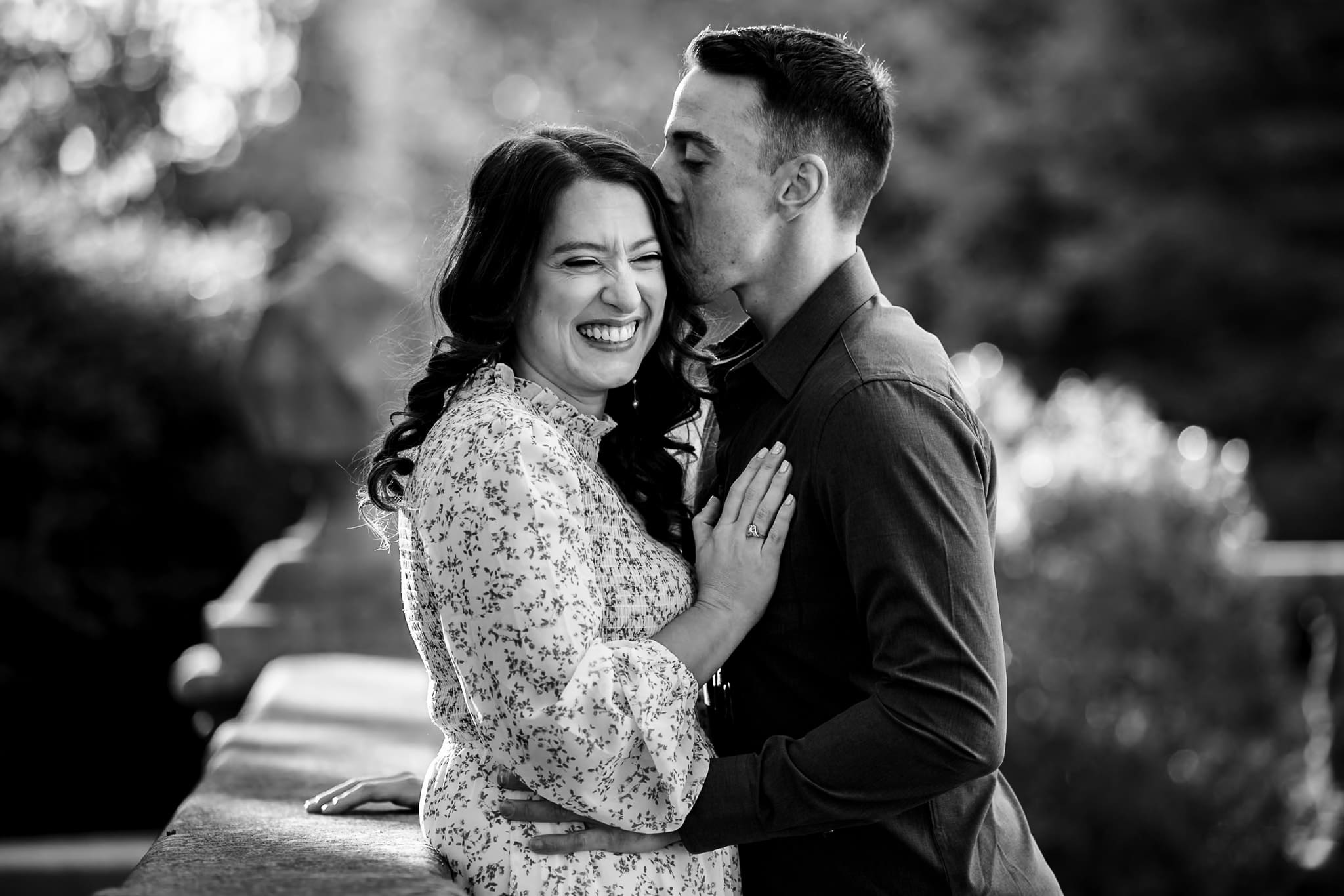 Groom-to-be kisses fiancée on the temple as she laughs during a romantic black and white engagement photo
