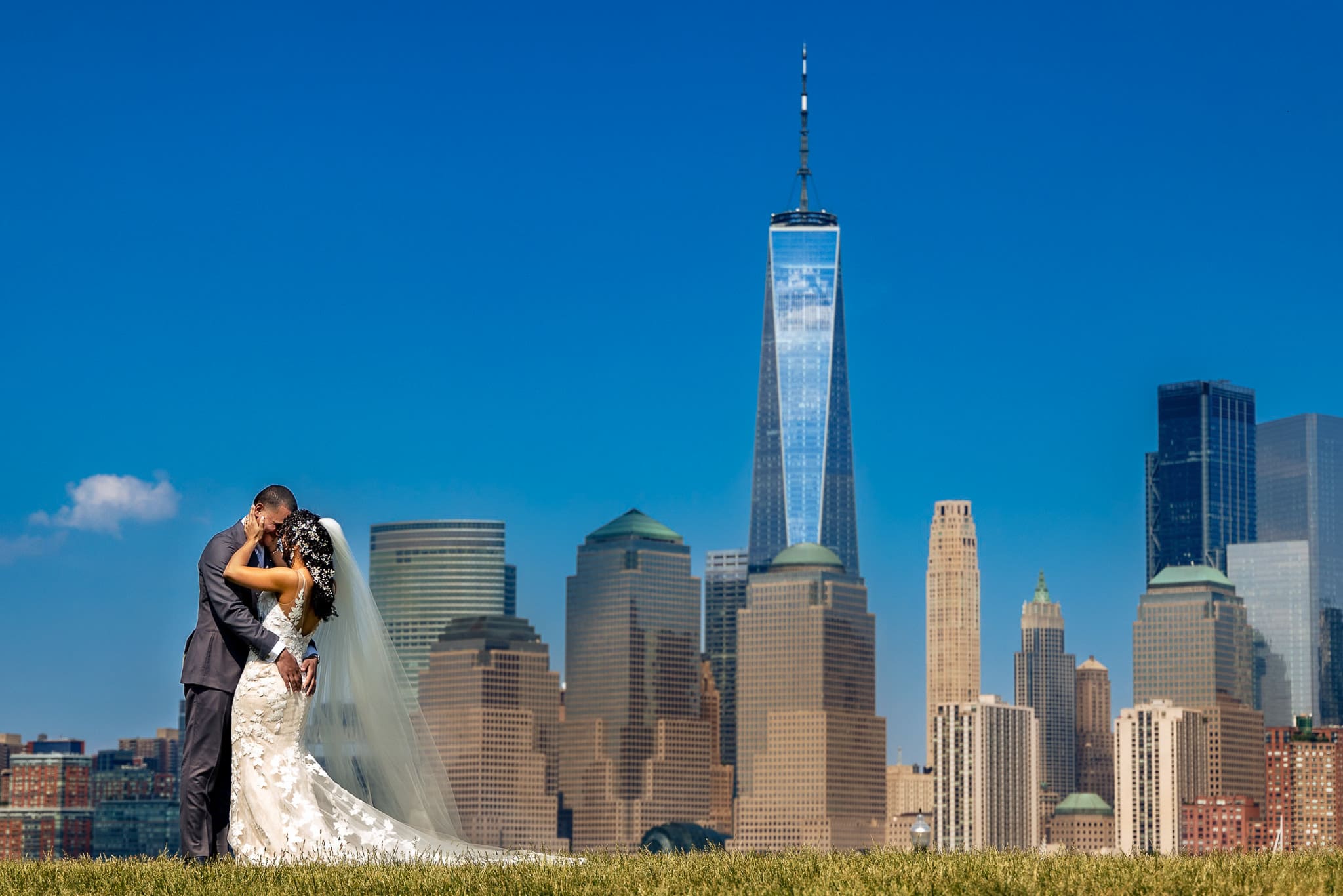 Bride and groom embracing with the New York City skyline and One World Trade Center in the background on a sunny day