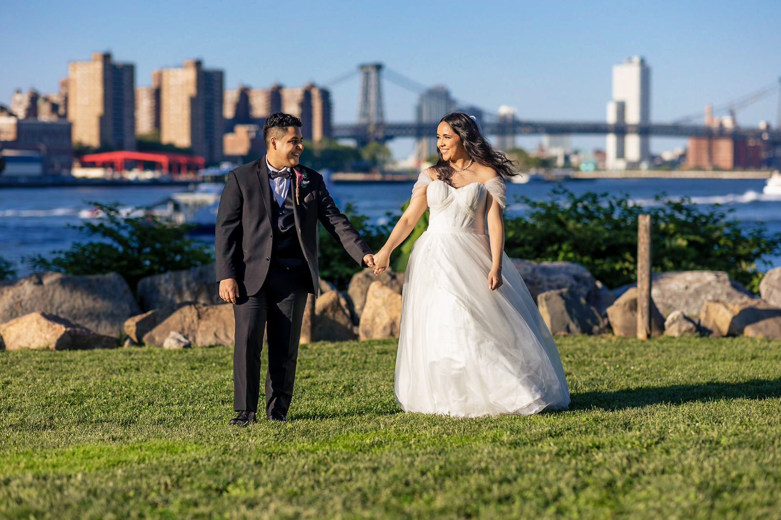 A bride and groom hold hands and smile outdoors on grass by a river, with city buildings and a bridge in the background under a clear blue sky—capturing the romantic charm often found at Brooklyn wedding venues.