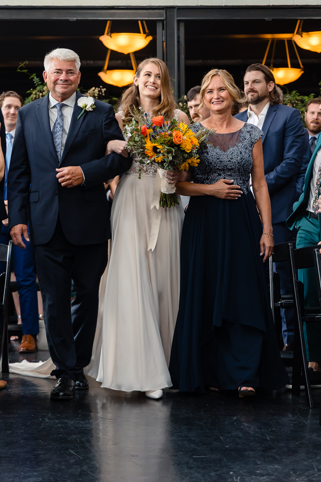 A bride in a white dress, holding a colorful bouquet, walks down the aisle smiling, accompanied by two smiling people in formal attire, surrounded by seated wedding guests.