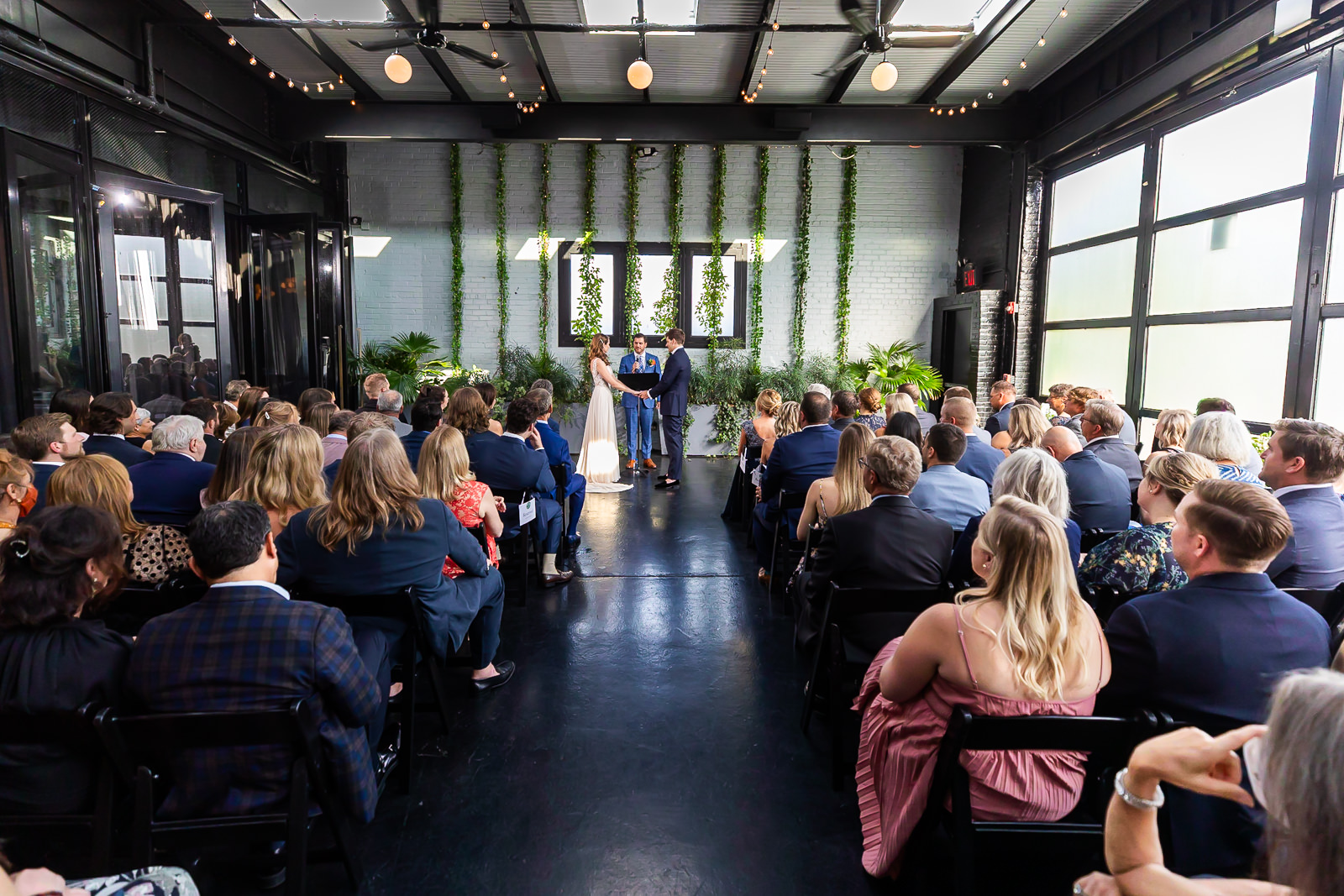 A wedding ceremony inside a modern venue with guests seated, a couple standing at the altar, and greenery decorating the white brick wall behind them.