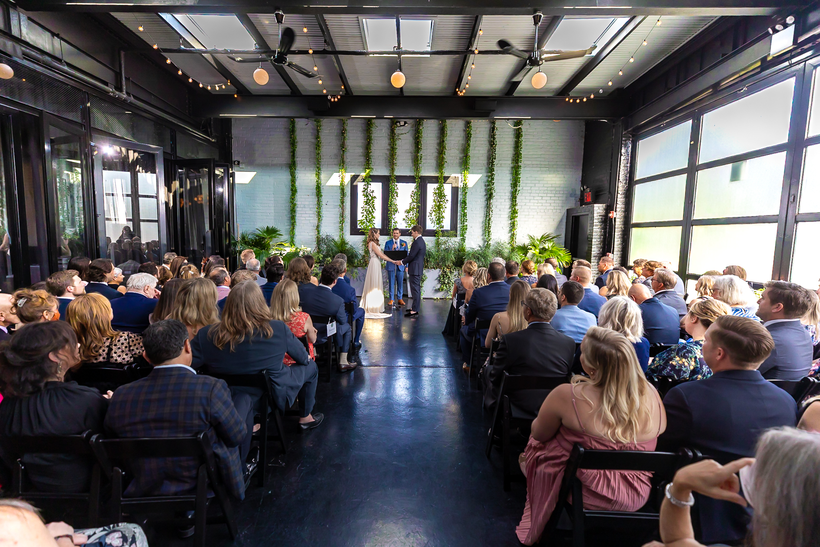 A couple stands at the altar in a modern, sunlit venue, surrounded by seated guests watching their wedding ceremony. Greenery decorates the white brick wall behind them.