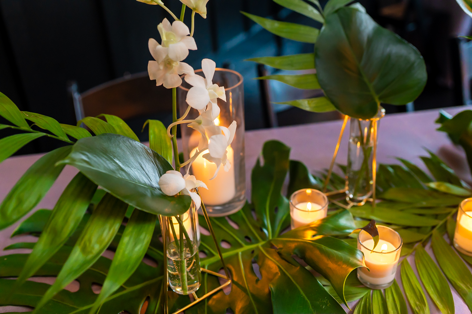 Green tropical leaves, white orchids in glass vases, and several lit candles are arranged decoratively on a table, creating an elegant and warm atmosphere.