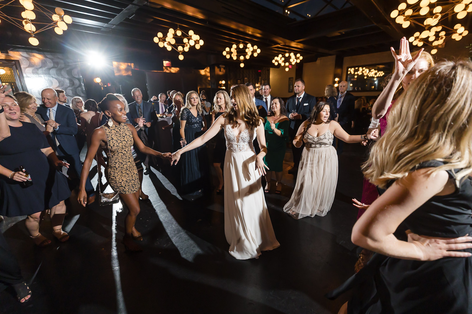A group of people dressed formally dance in a circle at an indoor event, smiling and laughing under warm, decorative lighting at a 501 Union wedding.