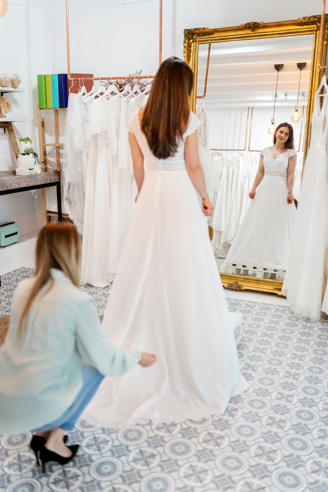 A bride tries on a white wedding dress in a boutique, looking at her reflection in a large gold-framed mirror while an assistant—recommended by top Wedding Planners in Brooklyn—adjusts the dress’s train on the patterned floor.