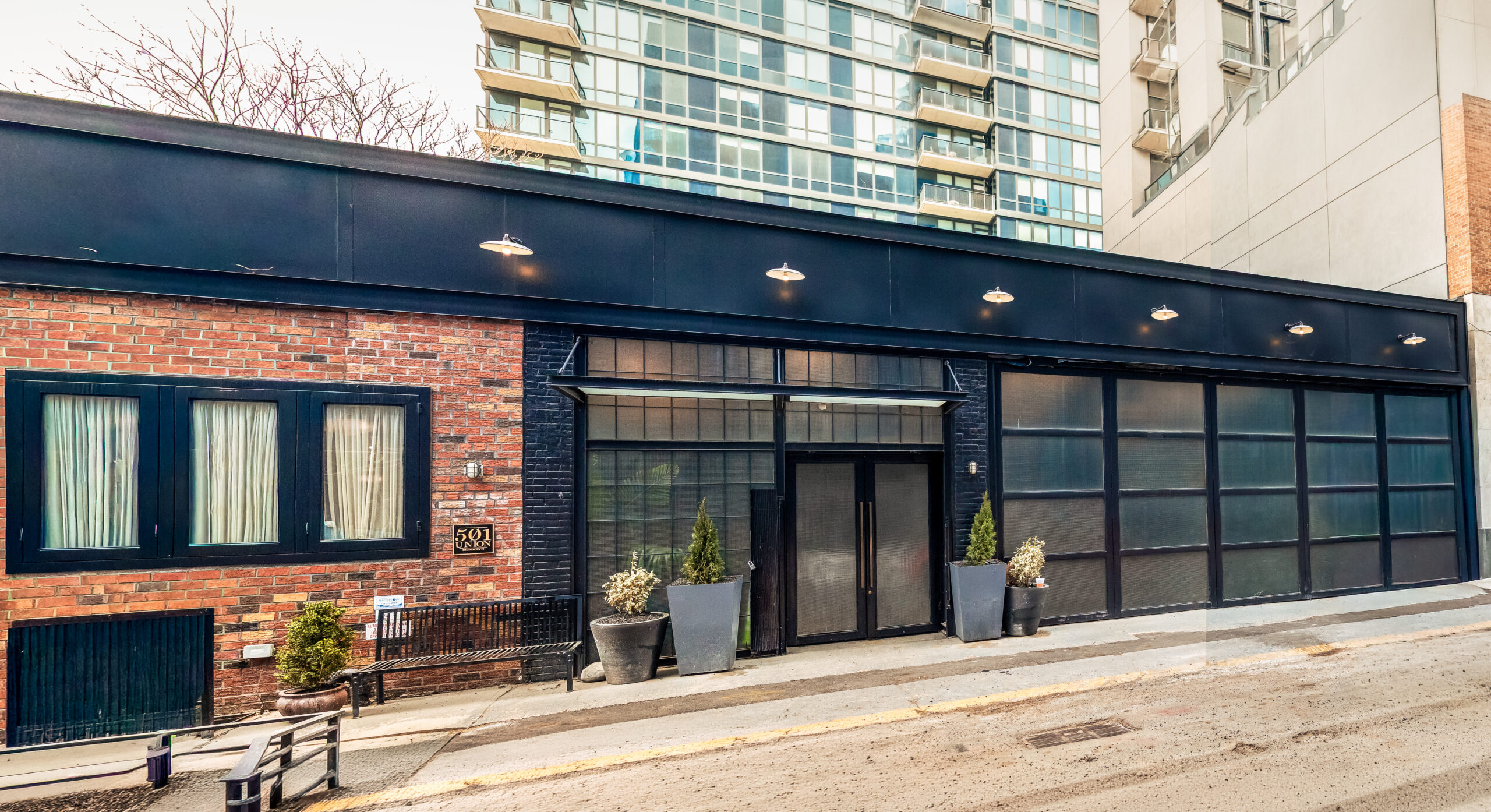 Modern building with large glass doors and windows, black trim, and brick accents. Several potted plants sit by the entrance. A tall residential building rises behind it. The street in front is sloped.