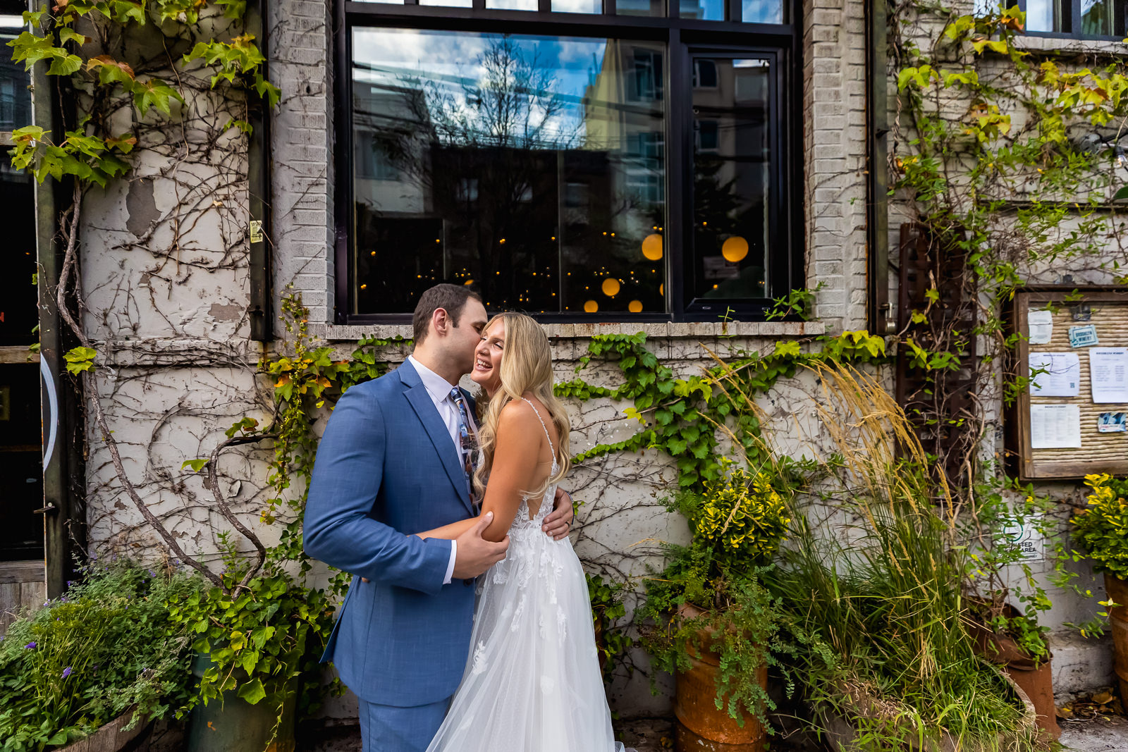 A bride and groom smile and embrace outdoors in front of a vine-covered brick wall and large window, with greenery and plants around them.