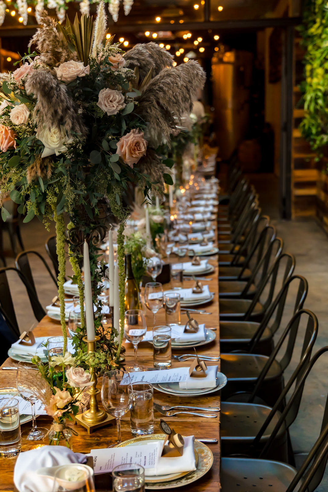 A long banquet table is set for a formal dinner with elegant floral centerpieces, candles, menus, plates, glasses, and utensils, surrounded by black chairs in a warmly lit room with string lights overhead.