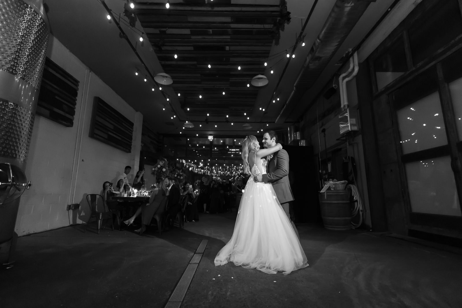 A bride and groom share their first dance in a dimly lit industrial venue, with guests seated at tables in the background and string lights overhead.