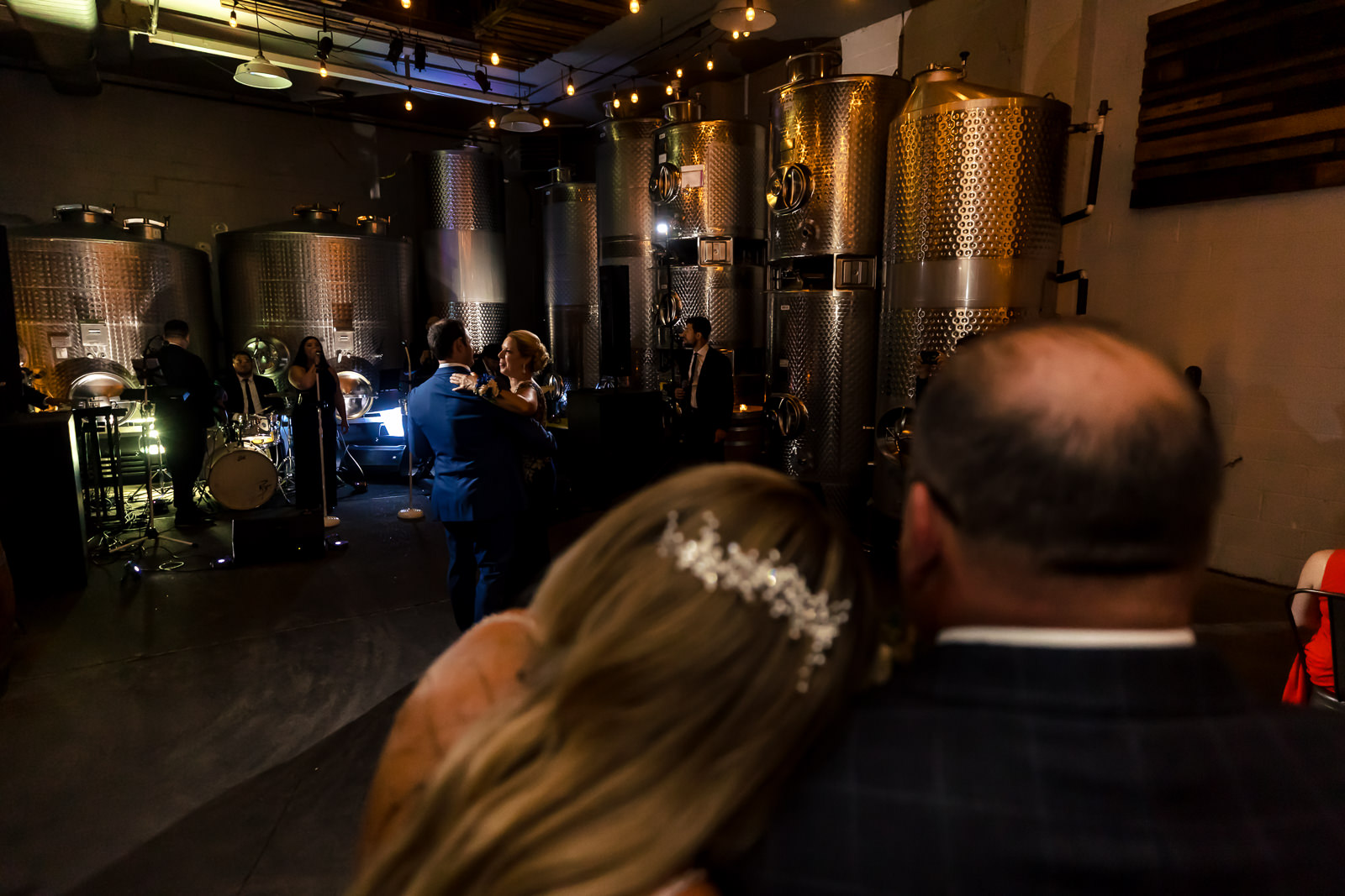 A couple sits close together watching people dance at a wedding reception inside a brewery, with large metal brewing tanks in the background, under warm string lights.