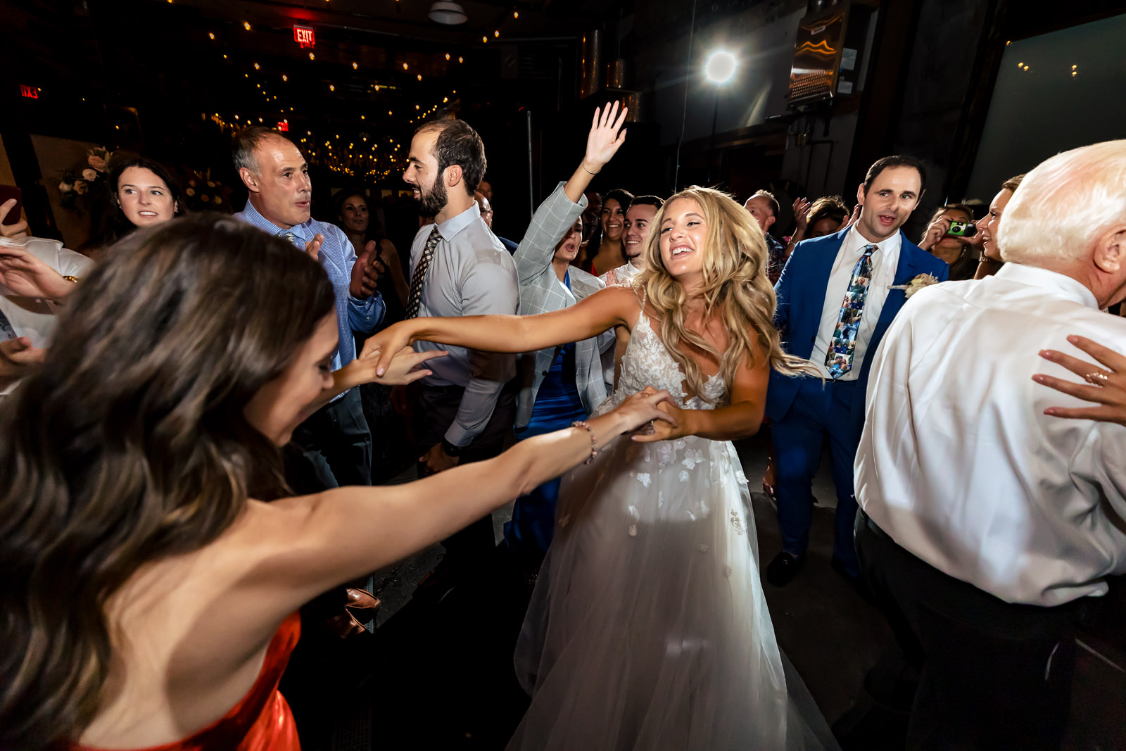 A bride in a white dress dances joyfully with guests at a lively wedding reception, surrounded by smiling people under string lights.