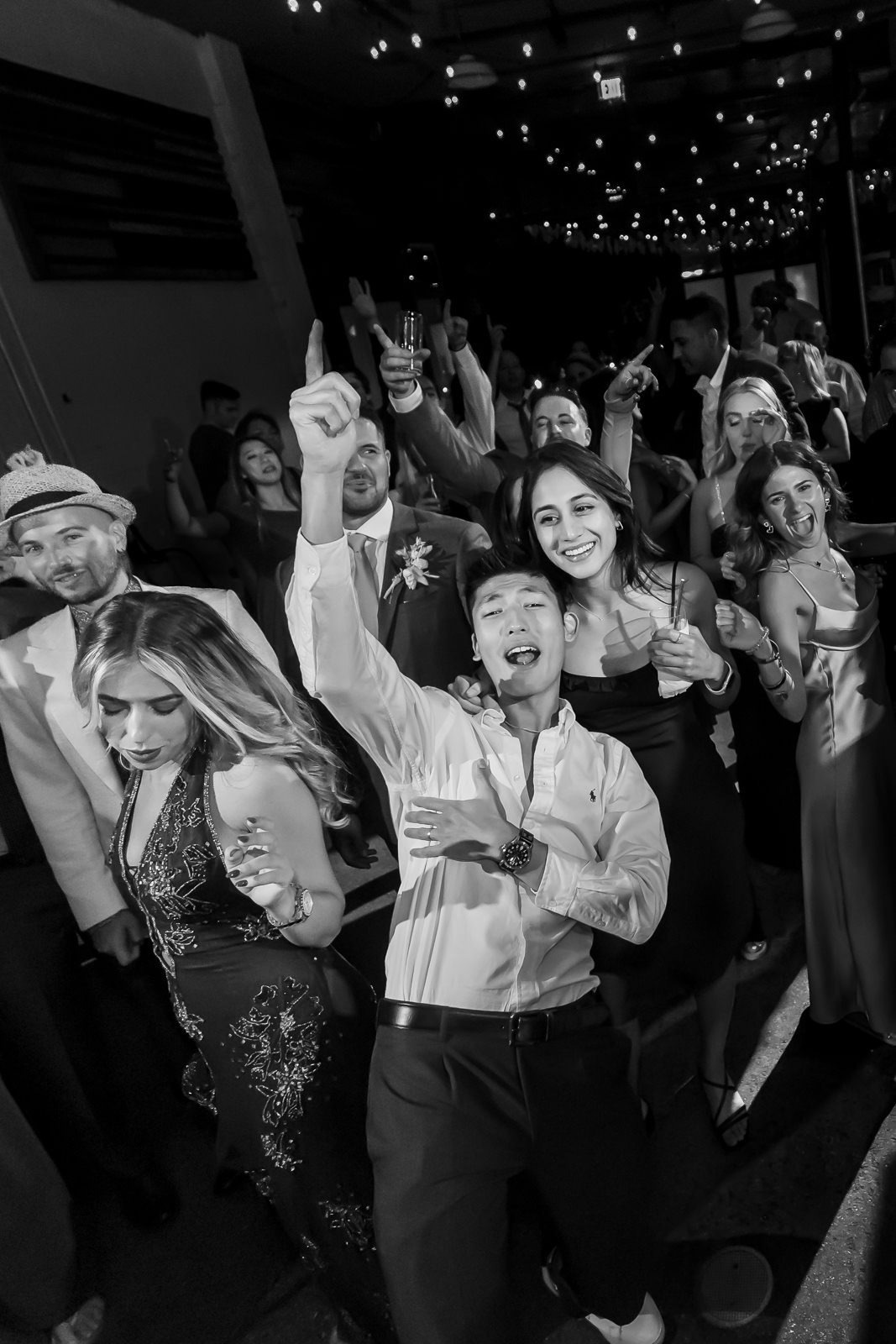 A group of people dancing and celebrating at a party; one person in front points upward and smiles, while others cheer and laugh under string lights.