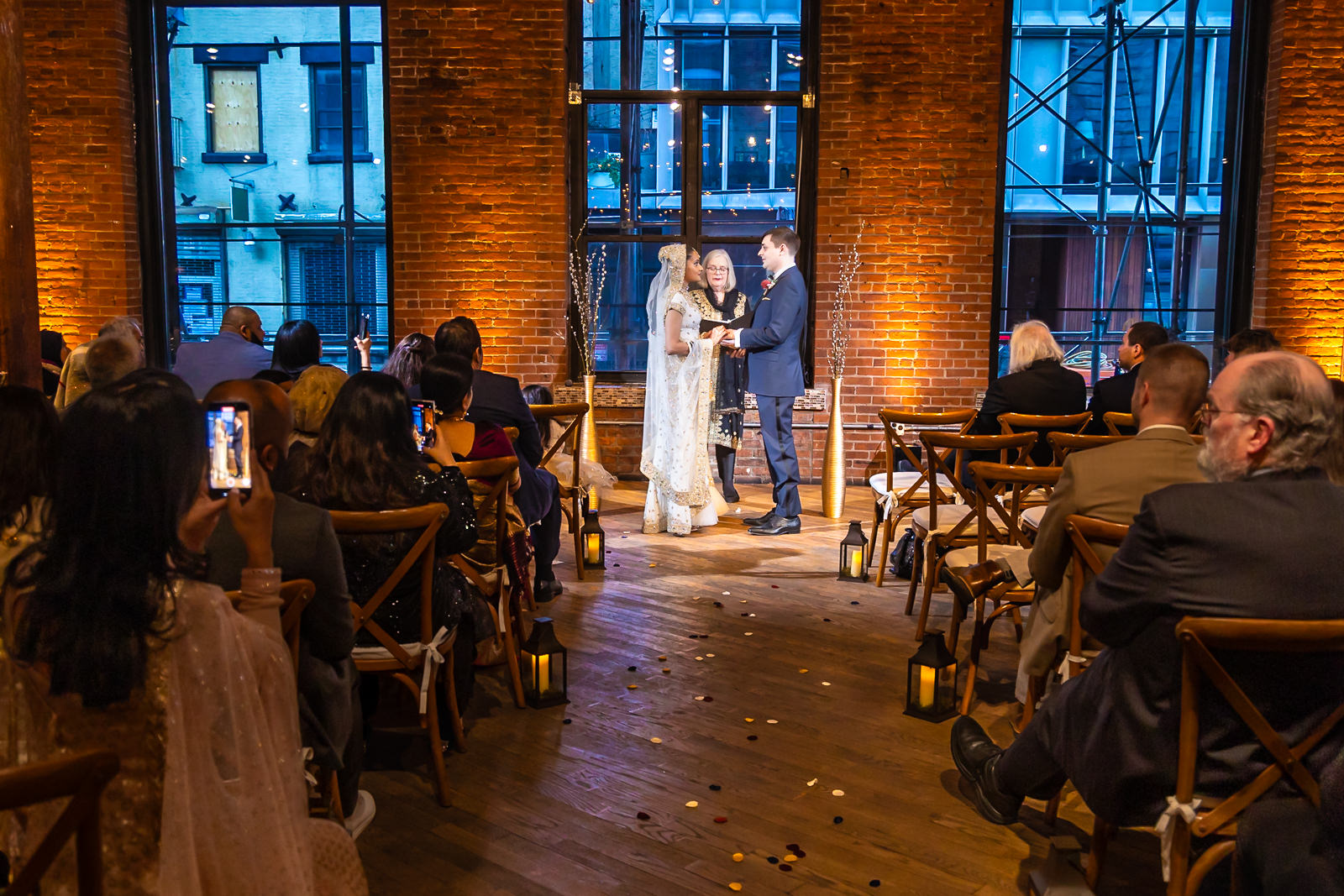 A couple stands facing each other at the altar during an indoor wedding ceremony, surrounded by seated guests and warm lighting from tall windows and exposed brick walls.