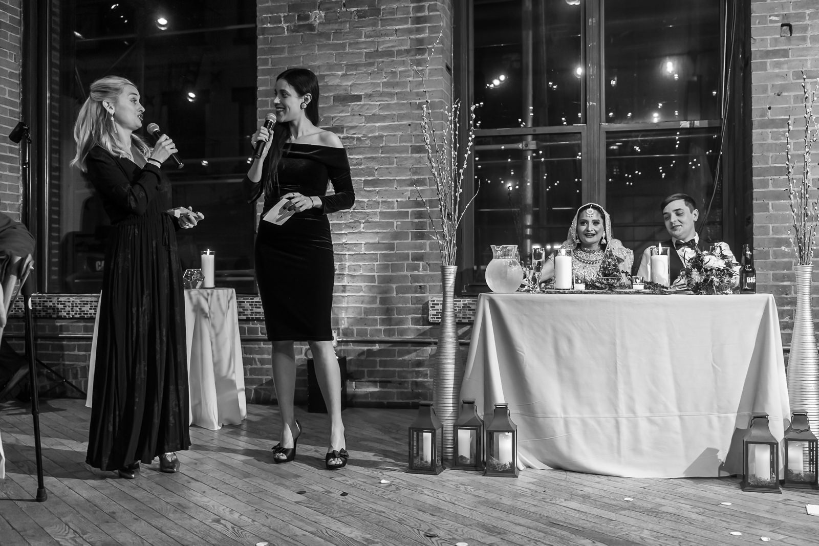 Two women stand speaking with microphones at a wedding reception, while a bride and groom sit at a decorated table nearby. Lanterns and candles decorate the wooden floor and table in a brick-walled venue.