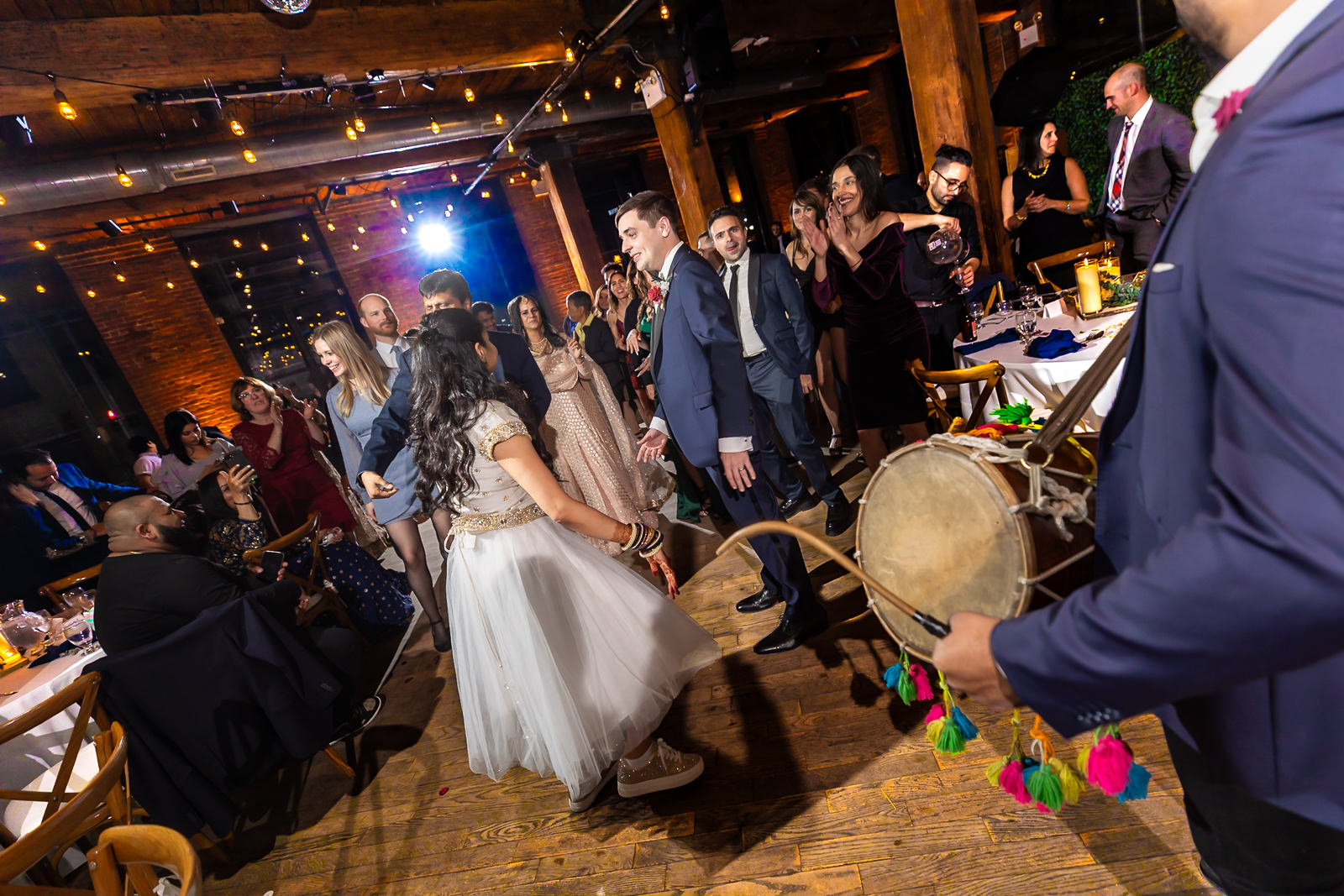 A group of people dance joyfully at an indoor celebration, with a musician playing a drum in the foreground. Warm lighting and exposed brick walls create a festive atmosphere.