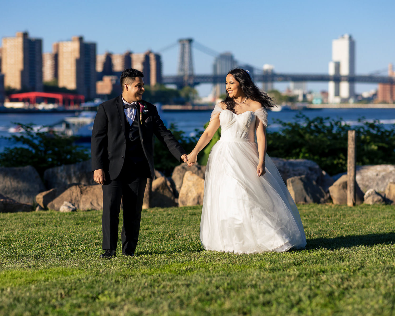 A bride and groom hold hands and smile at each other while standing on grass, with a river, city buildings, and a bridge in the background on a sunny day—capturing a perfect moment in their wedding day timeline.