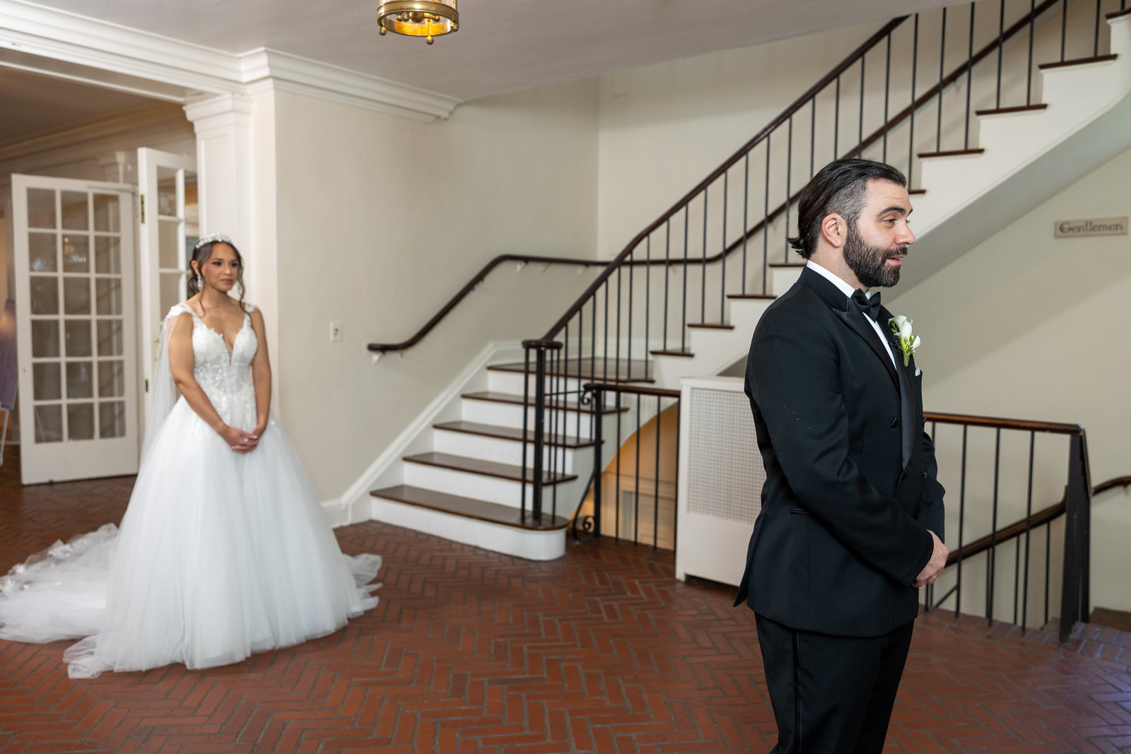 A bride in a white gown stands behind a groom in a black suit, who faces away from her, capturing the anticipation of their wedding first look in a room with brick flooring and a staircase.