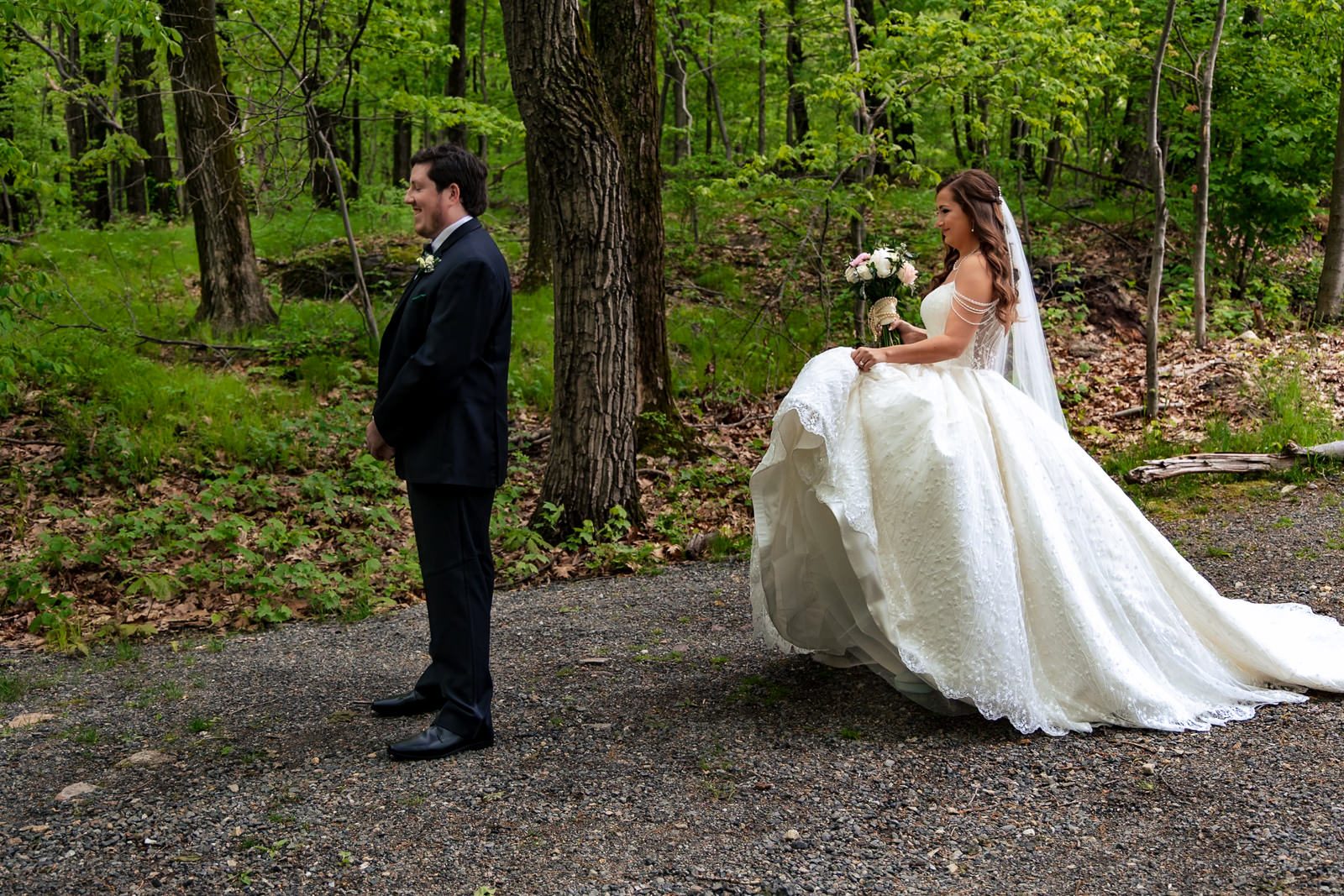 A bride in a white gown walks up behind her groom standing on a forest path, preparing for their magical wedding first look amid lush green trees.