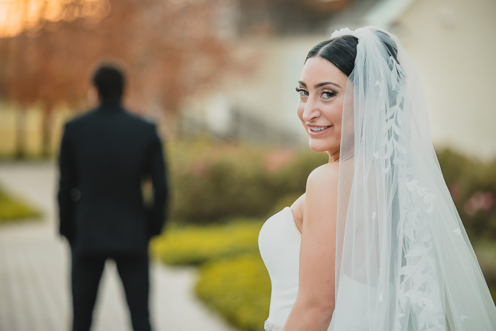 A bride in a white dress and veil smiles at the camera outdoors, savoring a joyful moment in her wedding day timeline, while a person in a black suit stands blurred in the background on a path.