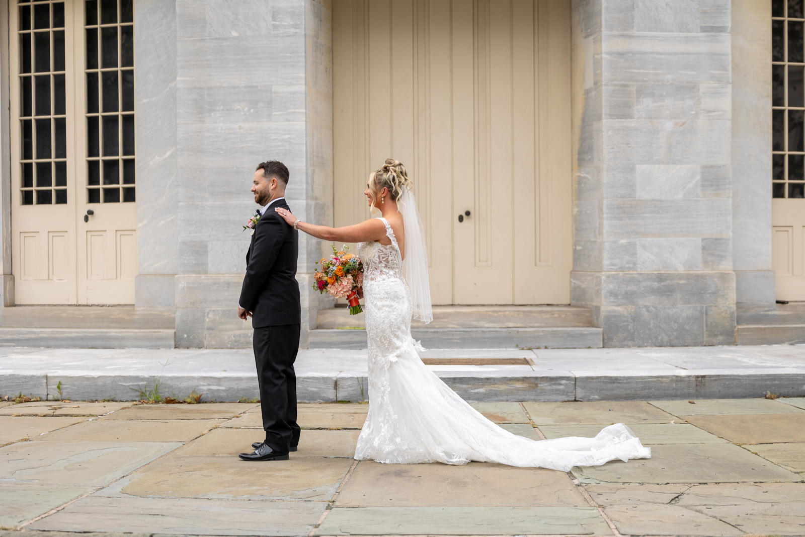 A bride in a white dress stands behind her groom in a black suit, gently touching his shoulder for their wedding first look, outside a building with tall windows and beige doors.