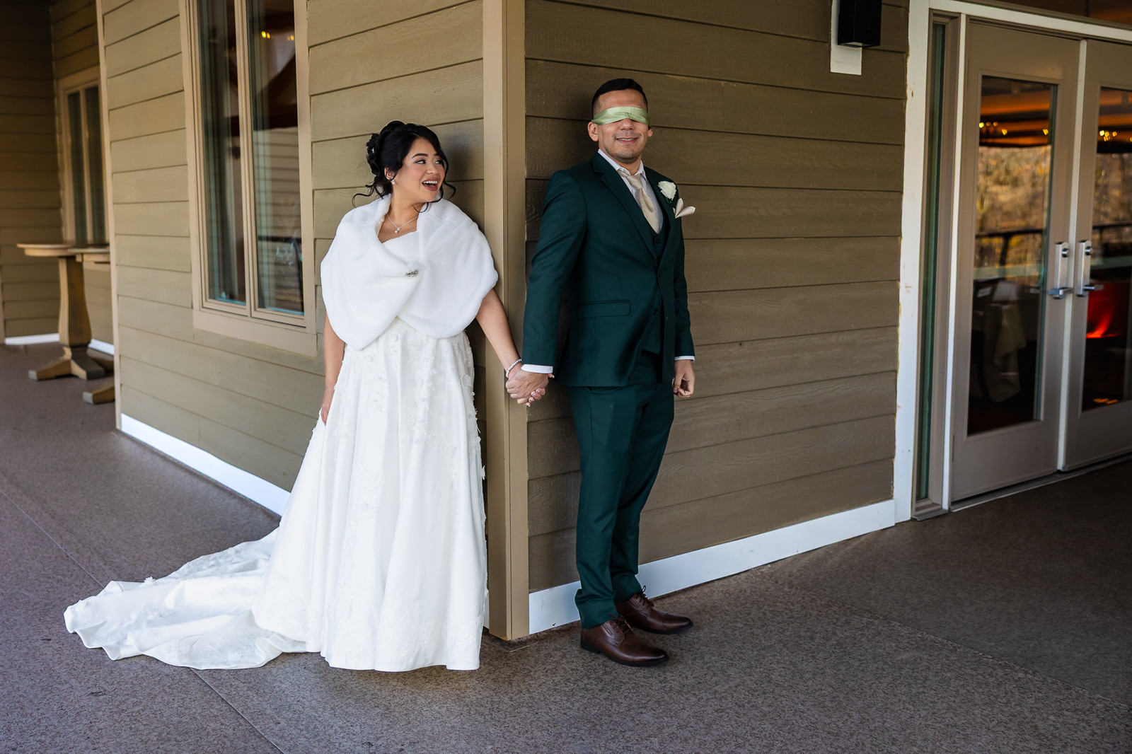 A bride in a white dress and a groom in a green suit, who is blindfolded, stand on opposite sides of a building corner holding hands and smiling during their wedding first look, just before their ceremony.