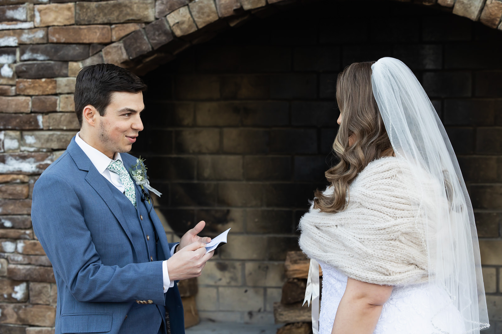A groom in a blue suit reads vows to a bride in a white dress and veil, standing in front of a rustic stone fireplace during their wedding first look. The bride faces away, while the groom looks at her, holding a piece of paper.