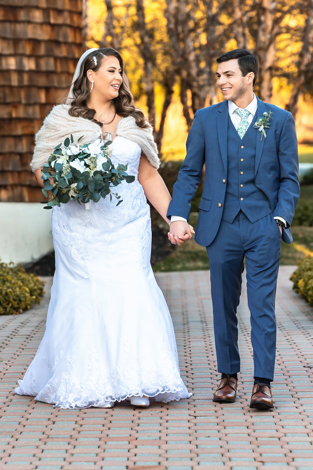 A bride in a white dress and shawl holds a bouquet while walking hand in hand with a smiling groom in a blue suit along a brick path outdoors, capturing the joy of their wedding first look.
