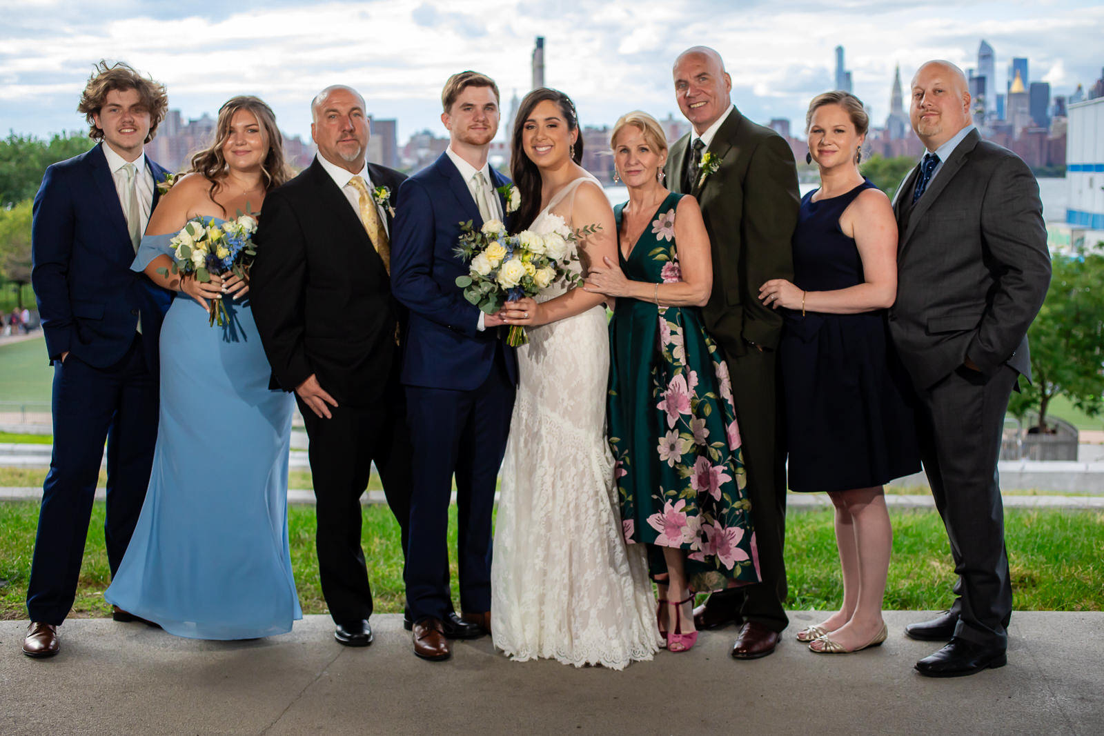 A wedding party of eight people, dressed formally, pose together outdoors. The bride and groom stand in the center, holding a bouquet, surrounded by family and friends, smiling at the camera as they celebrate each special moment on their wedding day timeline. A city skyline is visible in the background.
