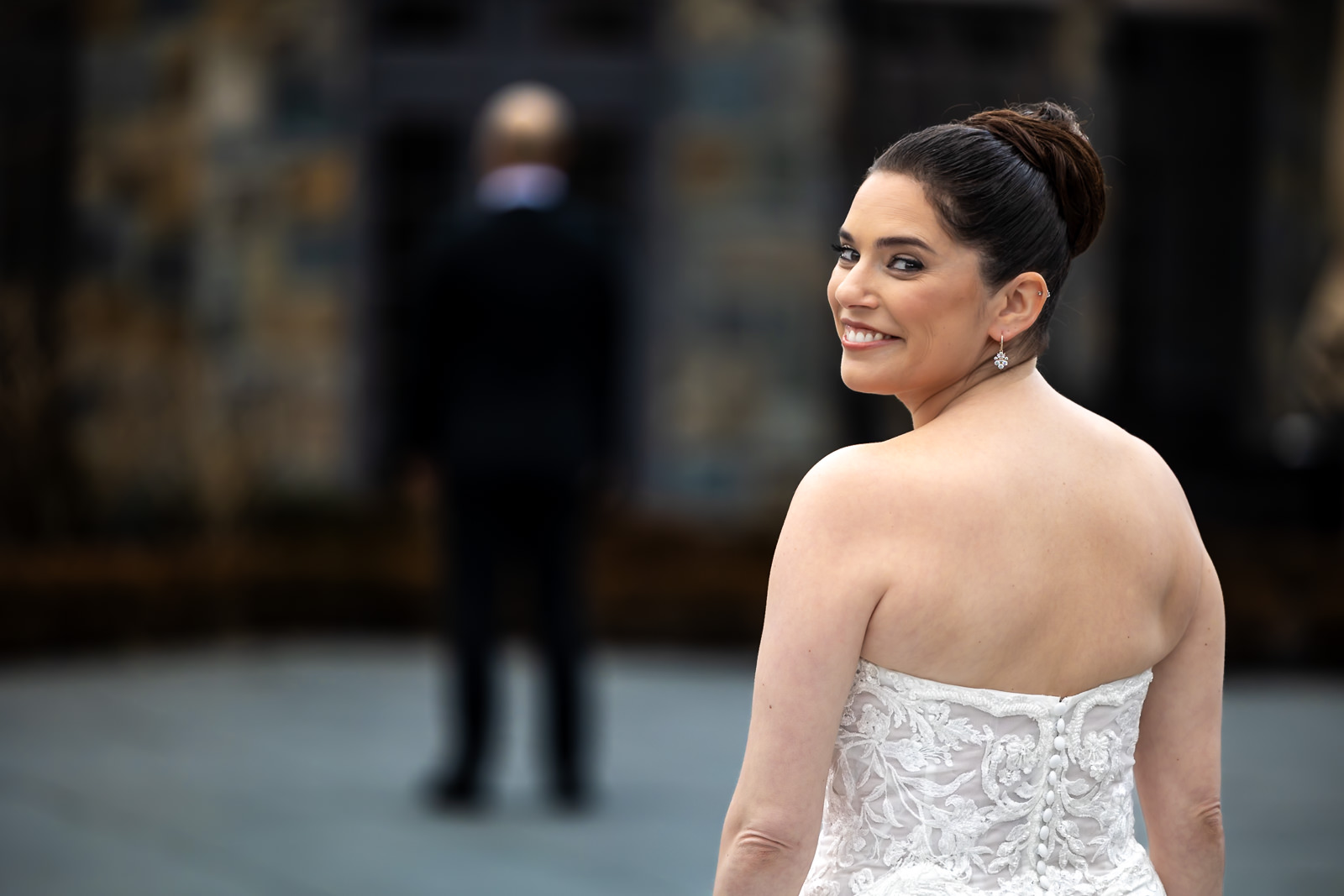 A bride in a strapless white gown smiles over her shoulder, facing the camera, capturing the excitement of a wedding first look, while a person in a dark suit stands blurred in the background.