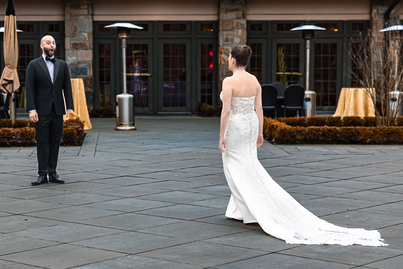 During a wedding first look, a bride in a white gown walks toward a man in a tuxedo, who looks surprised and excited. They are outside on a stone patio, with tables and heaters in the background.