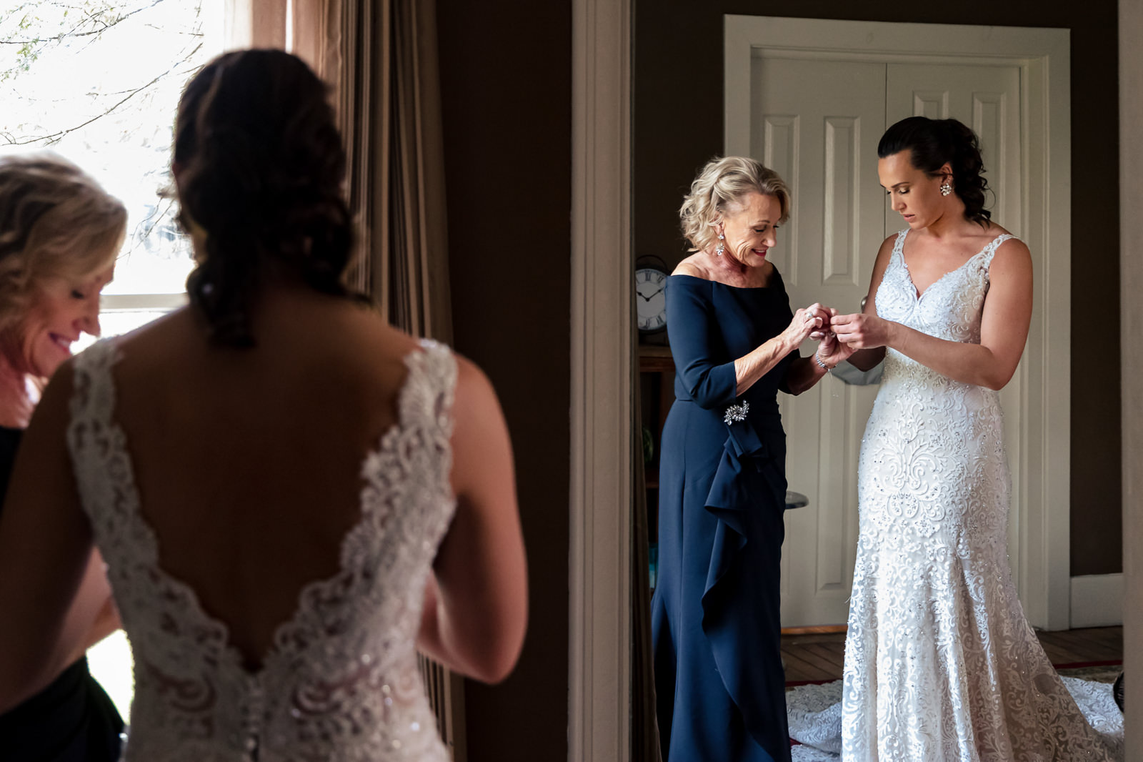 A bride in a lace gown stands with an older woman in a navy dress, sharing an intimate moment, as sunlight streams through a nearby window—one of those quiet pauses that make up the wedding day timeline. Another woman is seen in the foreground’s reflection, adding depth to this tender scene.