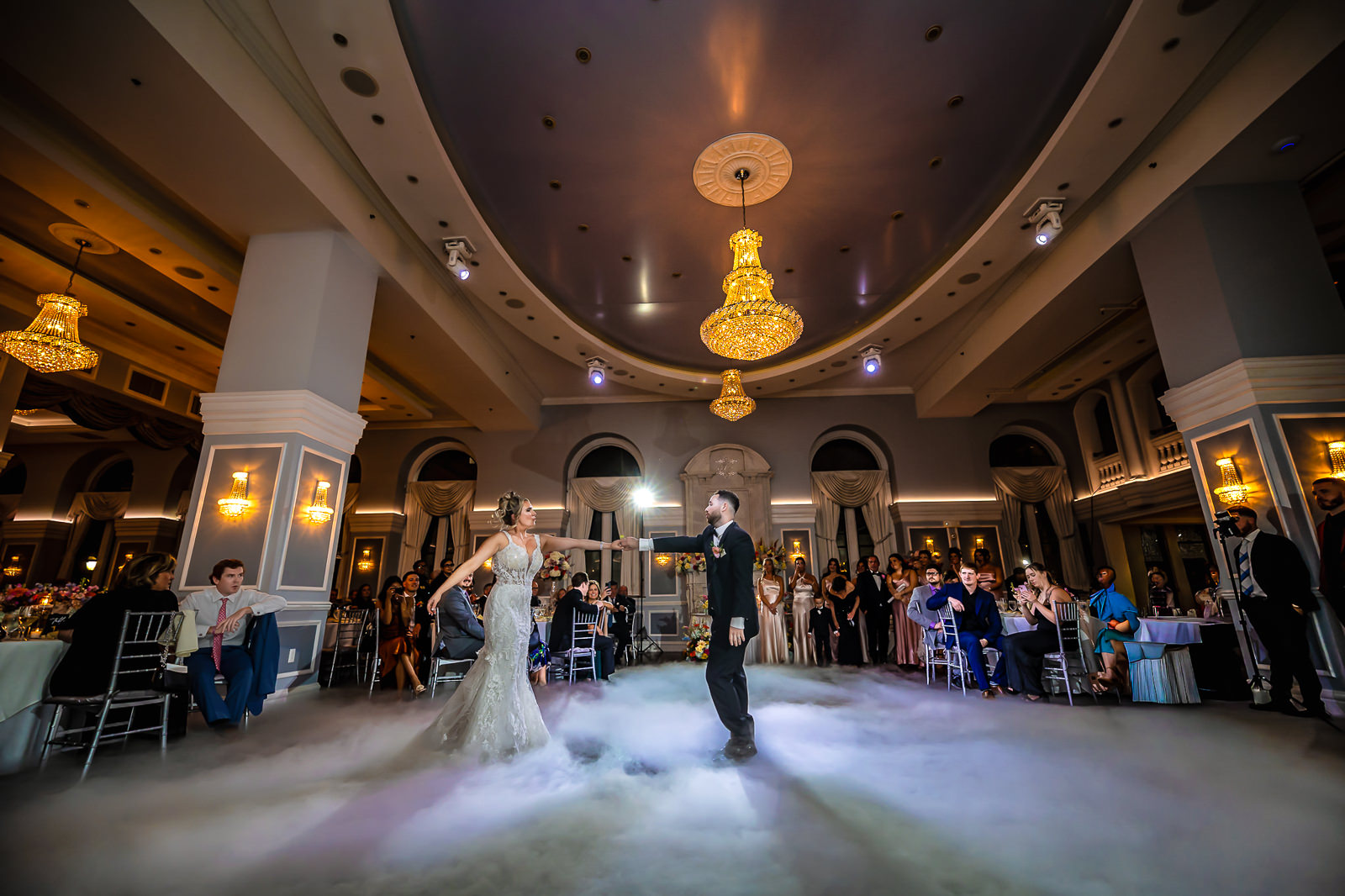 A bride and groom dance on a fog-covered floor in an elegant ballroom, surrounded by seated and standing guests. Chandeliers hang from the ceiling, adding a warm glow to the celebration—a magical moment perfectly placed in their wedding day timeline.