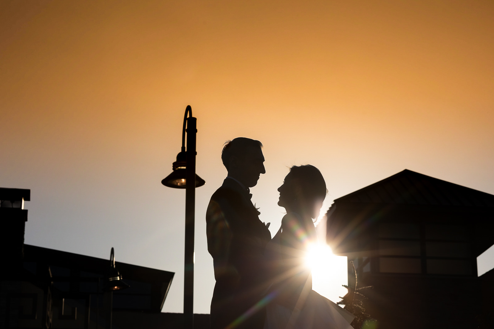 Silhouette of a couple embracing outdoors during sunset, with the sun shining between them and modern buildings in the background—a romantic moment that could be beautifully captured on your wedding day timeline.